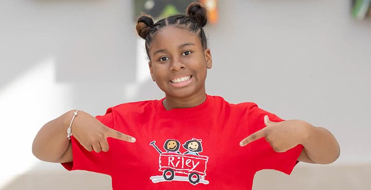 Girl wearing a red shirt with the Riley wagon logo, pointing to the logo on her shirt with both hands. 